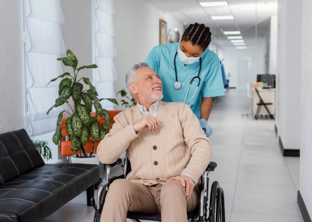 A care worker in scrubs pushing a smiling man in a wheelchair along a care home corridor