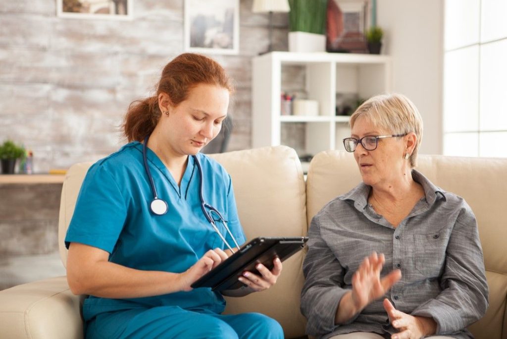 A nurse with a tablet listening to an older woman on a sofa