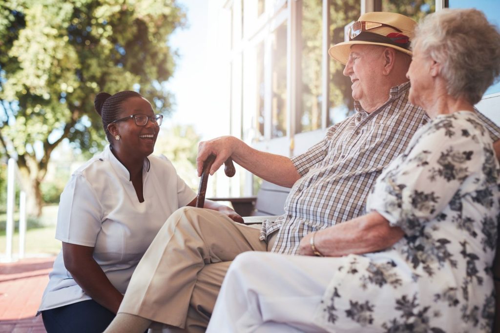 A carer crouching beside an older couple on a garden bench, all three laughing