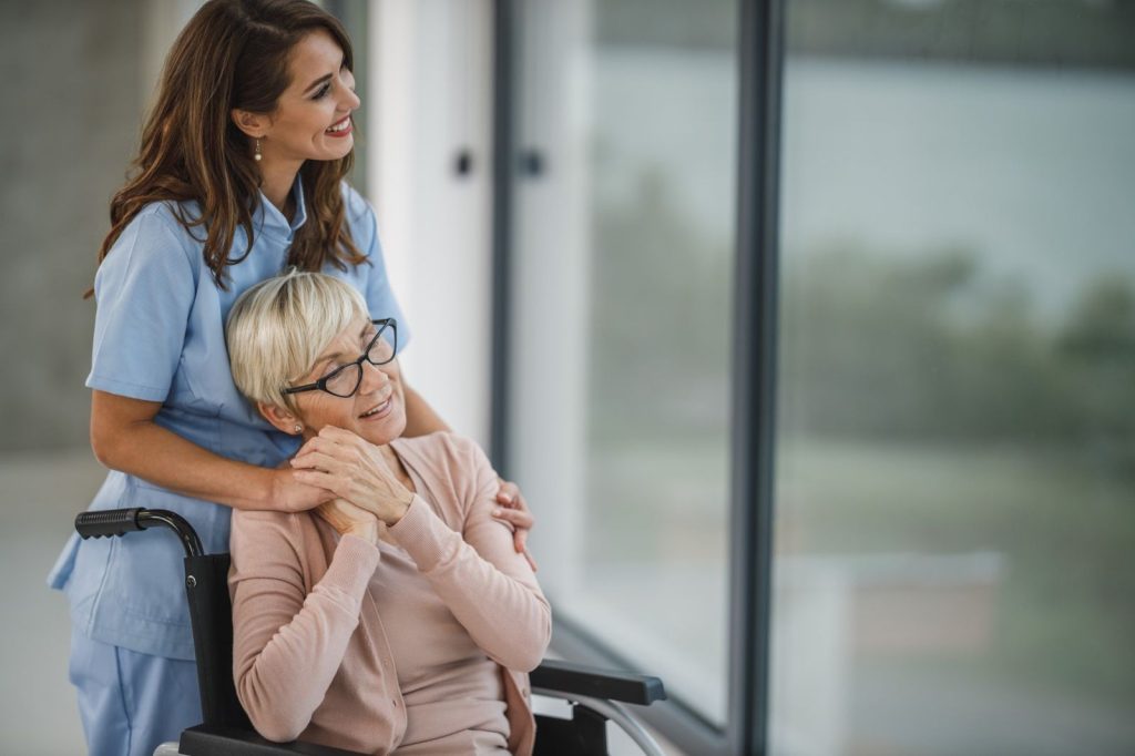 A carer with her arms around a woman in a wheelchair, both smiling and looking out of a window