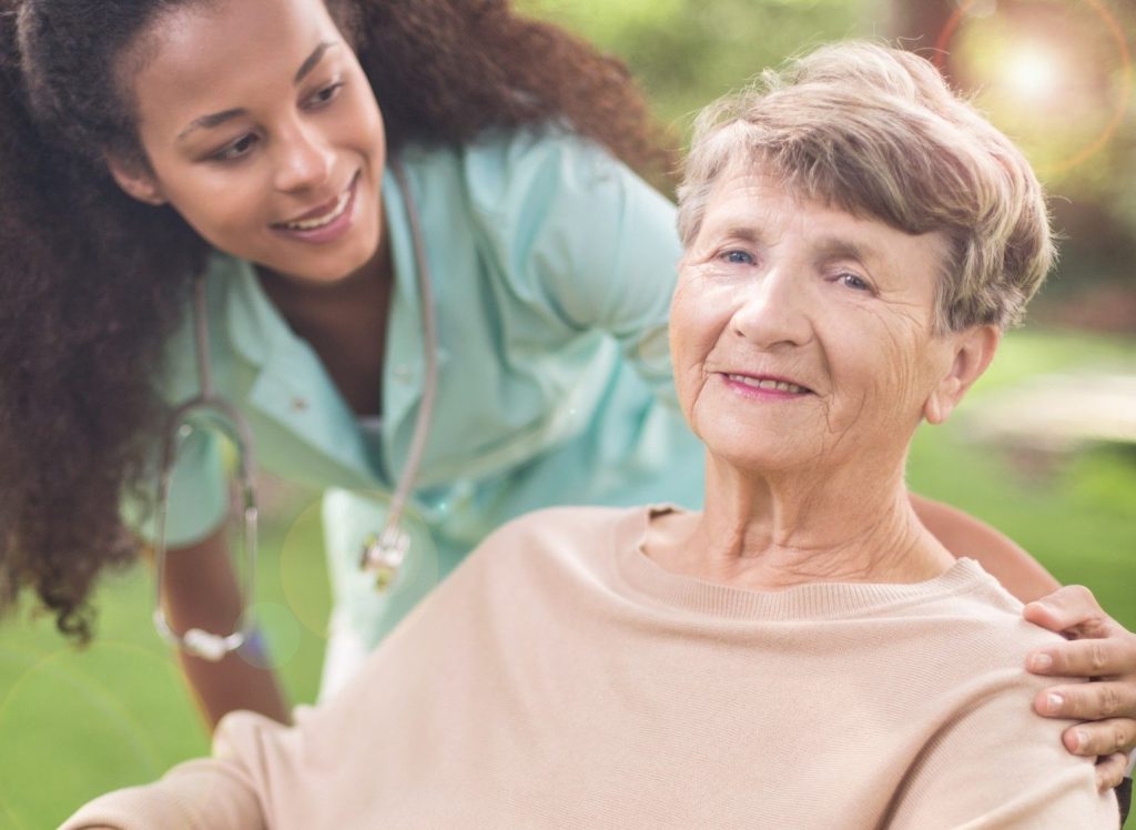 A carer with her hand on the shoulder of a smiling older woman in a garden