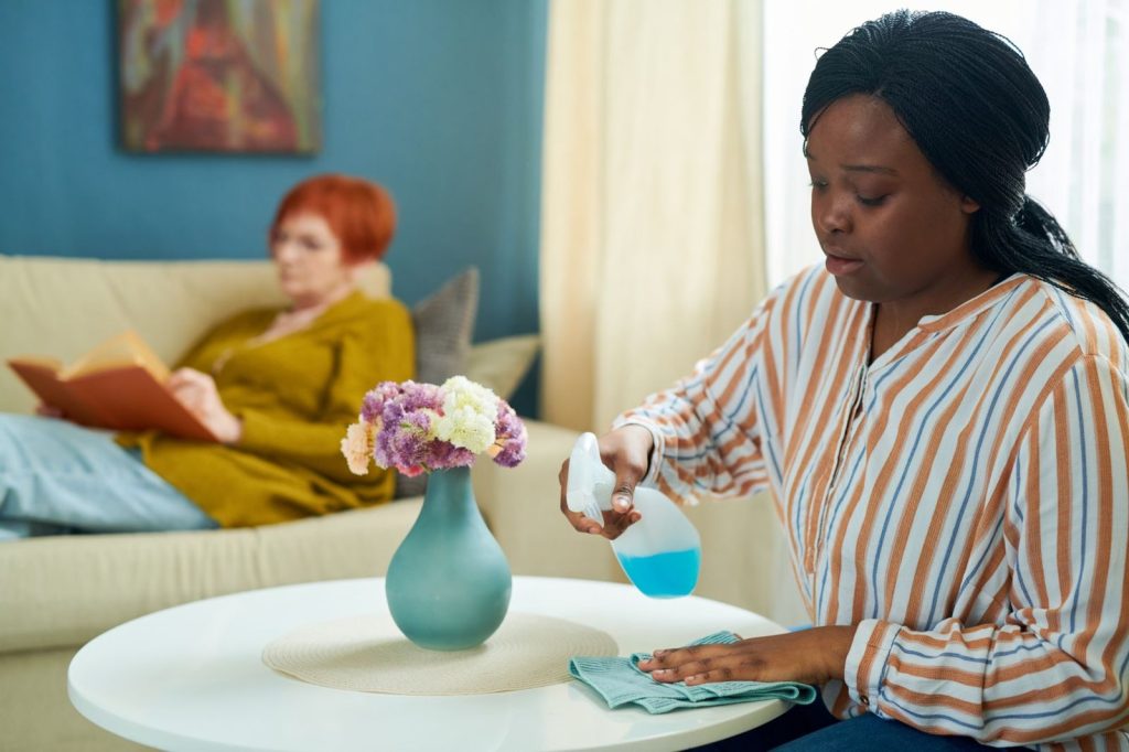 A home help wiping a table while an older woman reads on the sofa
