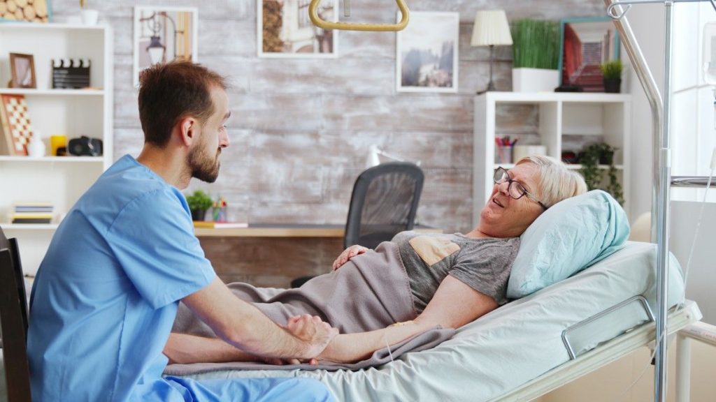 A nurse holding the hand of a woman resting in bed and chatting with her