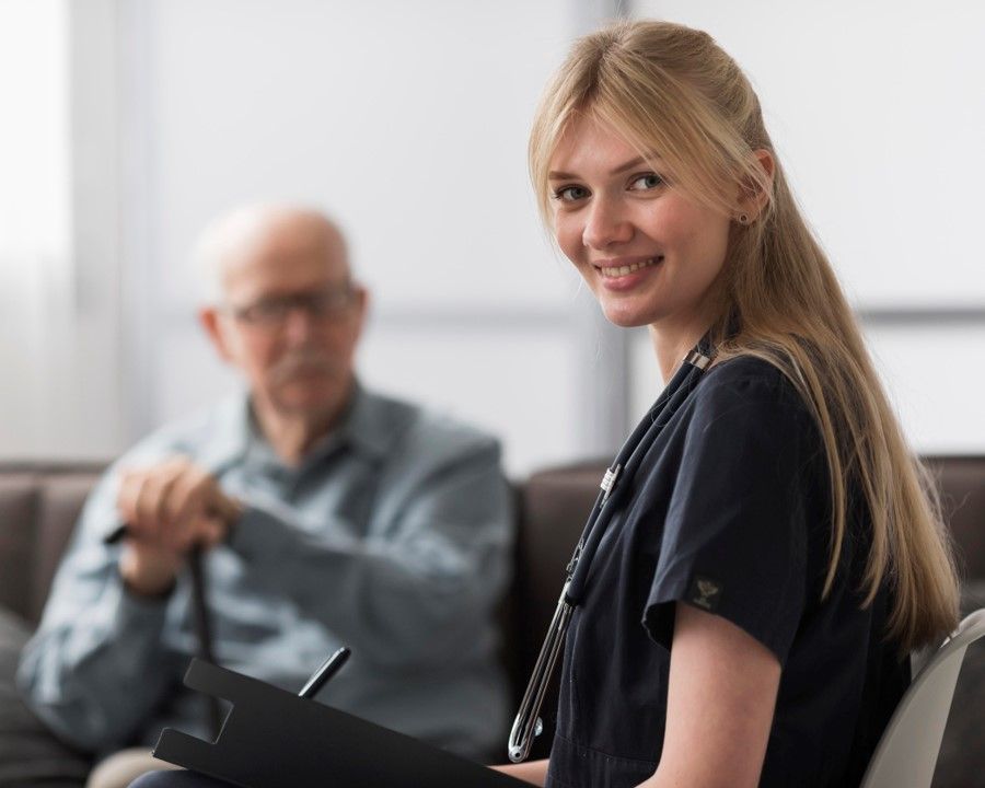 A smiling nurse holding a clipboard, with an older man sitting behind her