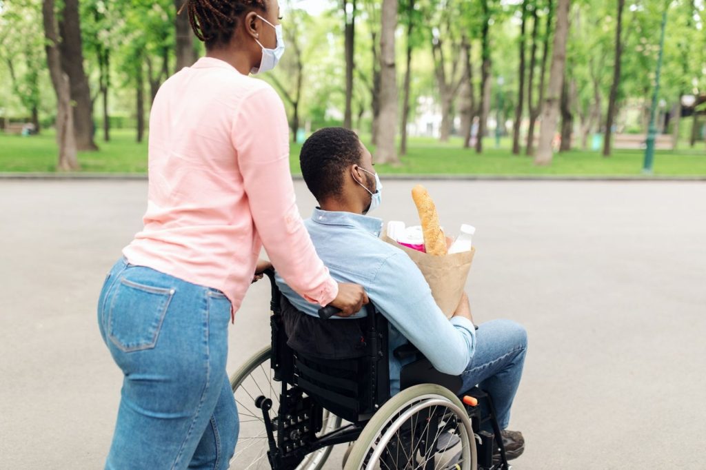 A woman pushing a man in a wheelchair through a park, with shopping on his lap